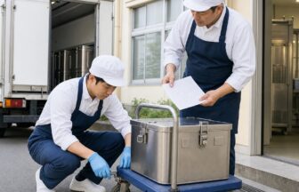 学校の受配口で先輩から給食容器と台車の確認方法を教わる新人配送員