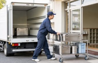 学校の受配口で密閉された給食容器を台車に載せて配送車から運ぶドライバー